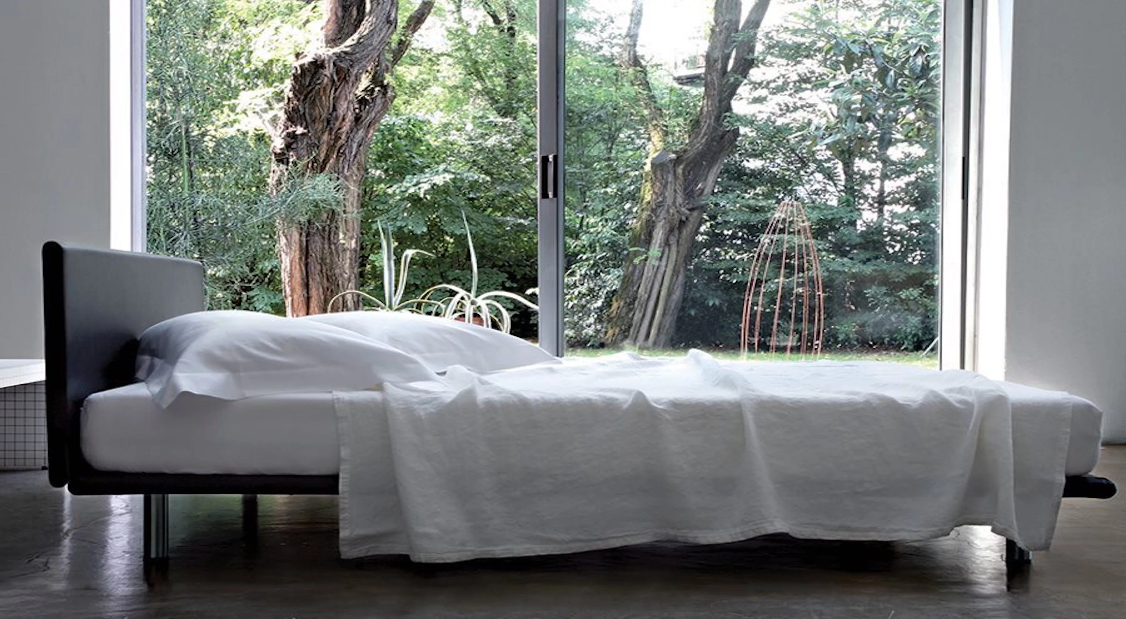 Ein modernes Bett von Milano in Weiß mit schwarzem Rahmen, vor einer großen Fensterfront mit Blick auf einen grünen Garten.