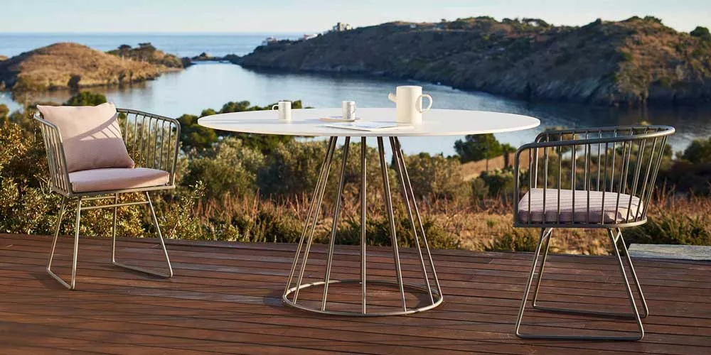 Bivaq Illa Essgruppe in Weiß und Metall auf Holzterrasse mit Meerblick, bestehend aus rundem Tisch und zwei Stühlen mit Kissen.