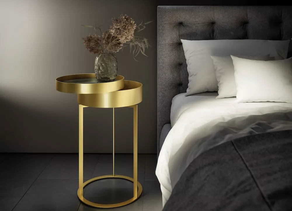 DRAENERT NIGHT Couchtisch goldfarben steht neben einem Bett mit grauem Kopfteil und weißen Kissen auf dem Tisch steht eine Vase mit getrockneten Blumen