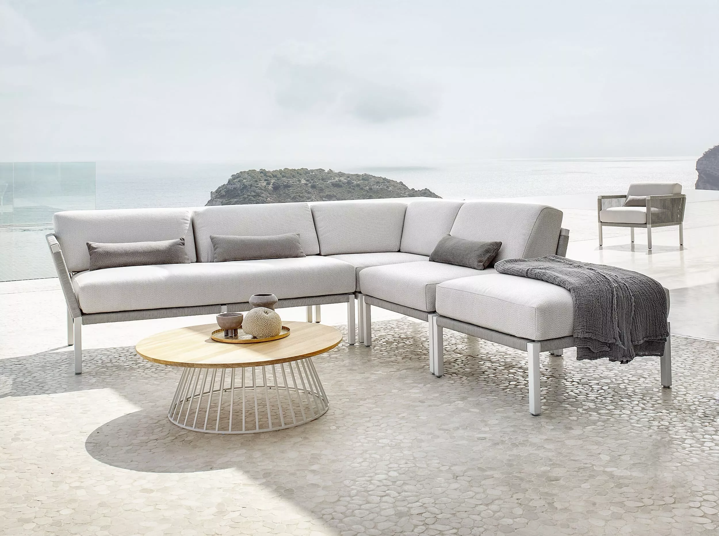Solpuri Club A Loungeecke in Weiß mit grauen Kissen und Decke auf einer Terrasse mit Meerblick und einem runden Holztisch im Vordergrund.