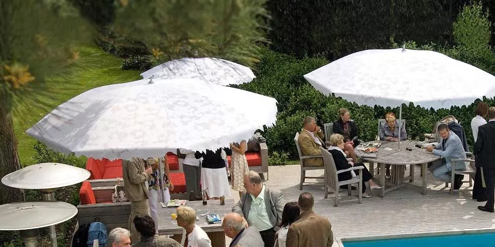 Personen entspannen sich unter weißen Sywawa Shadylace Sonnenschirmen im Garten, umgeben von grüner Hecke und Holzmöbeln.