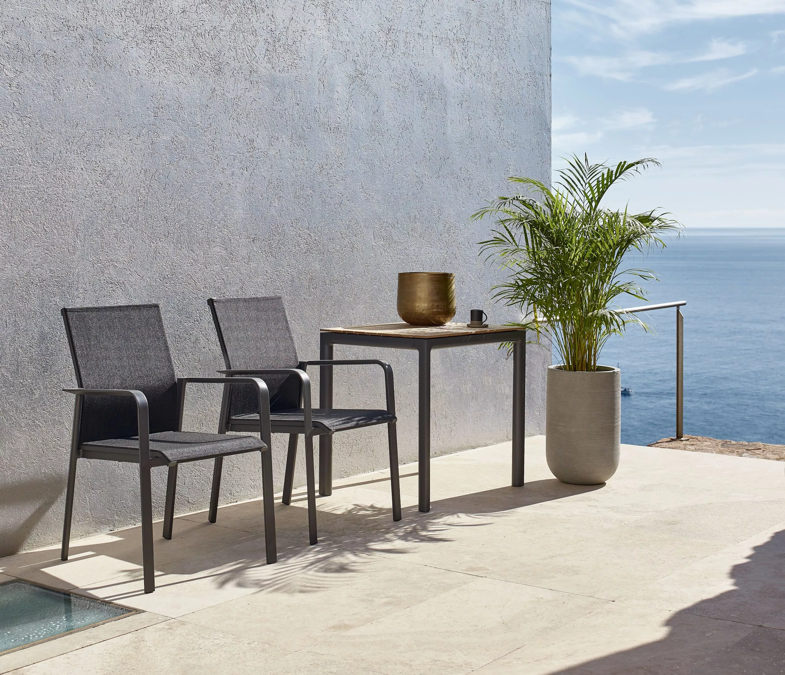 Zwei Solpuri Breeze Stühle und ein Tisch stehen auf einer Terrasse mit Blick auf das Meer neben einer großen Pflanze in einem Topf.