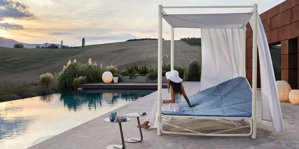 Frau in weißem Kleid und Hut entspannt auf der blauen Roberti Atollo Outdoor-Loungegruppe neben einem Pool bei Sonnenuntergang.