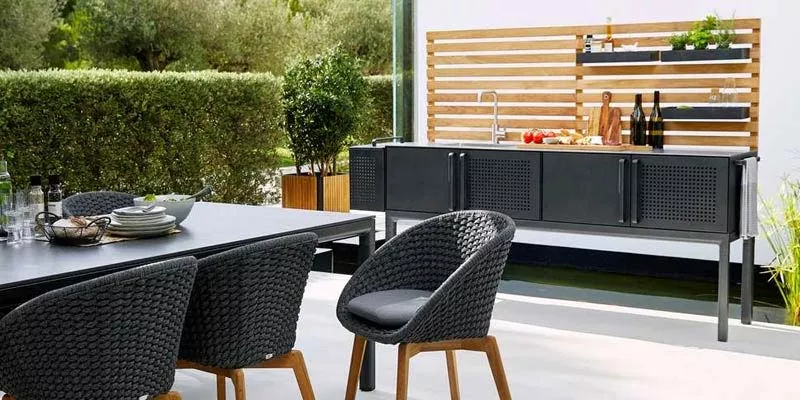 Cane-line Drop Outdoor-Küche in Schwarz auf Terrasse mit Tisch und Stühlen, umgeben von grüner Hecke und Pflanzen im Hintergrund.