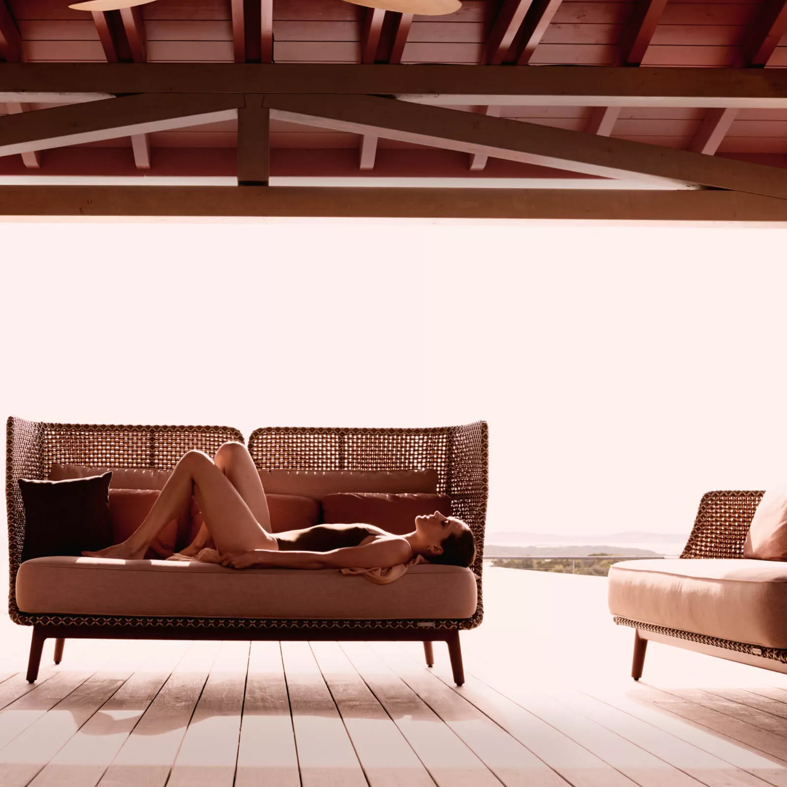 Eine Frau liegt entspannt auf einem Dedon Mbarq 3-Sitzer Sofa mit hoher Rückenlehne auf einer überdachten Terrasse mit Blick auf die Landschaft.