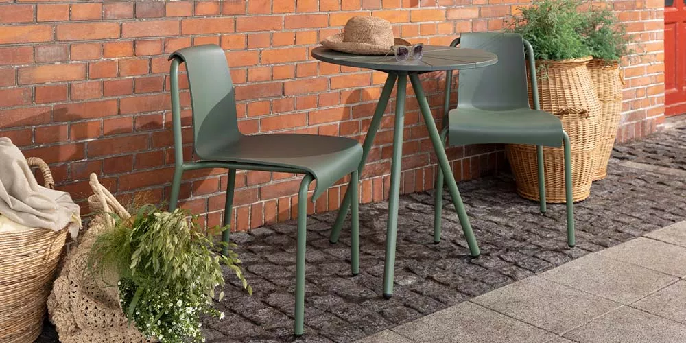 Zwei grüne Houe Nami Gartenstühle und ein runder Tisch auf Kopfsteinpflaster vor einer Backsteinmauer, dekoriert mit Pflanzen.