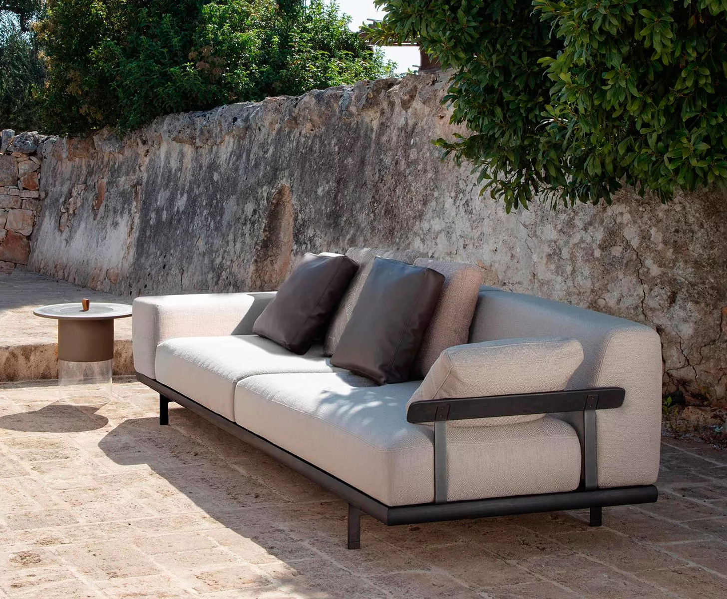 Talenti Allure Gartenmöbel Loungesofa in Beige auf einer Terrasse mit Steinmauer und Pflanzen im Hintergrund, seitliche Perspektive.