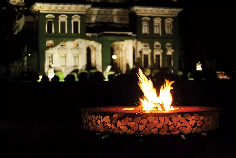 AK47 Zero Feuerstelle 200 cm Gartenkamin brennend im Garten vor einem beleuchteten Herrenhaus bei Nacht