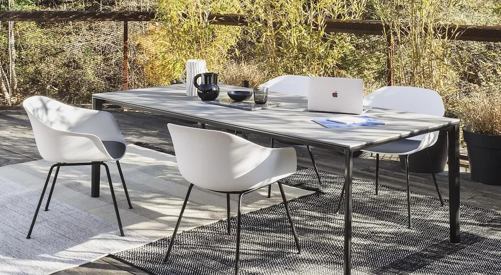 Vier weiße Stühle und ein grauer Tisch von Fast Aria auf einer Terrasse mit Laptop und Kaffeekanne, umgeben von Pflanzen.