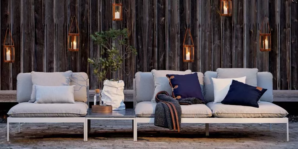 Skargaarden Bönan Sofa in Beige auf einer Terrasse mit Holzwand und Laternen im Hintergrund, dekoriert mit Kissen und Decke.