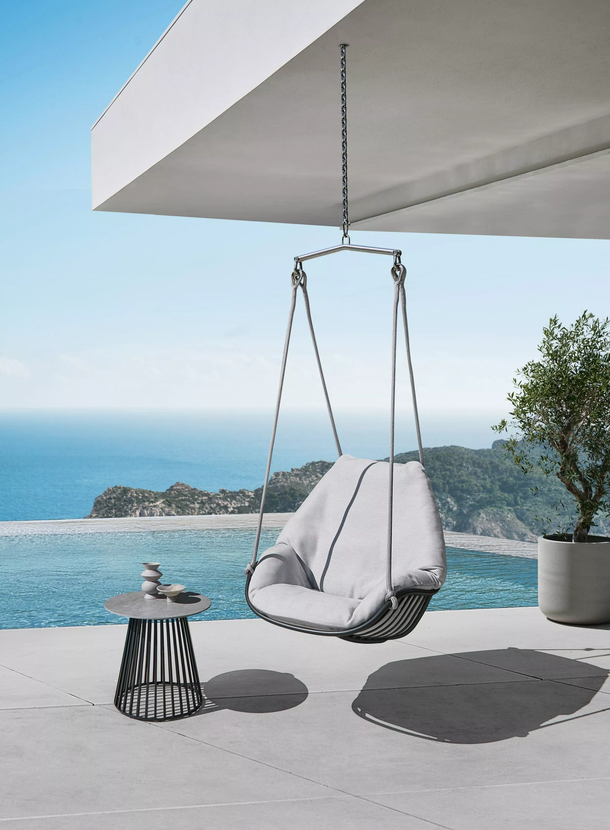 Solpuri Chill-A Hängesessel in modernem Design auf einer Terrasse mit Meerblick und einem Beistelltisch mit Dekorationen Moodbild