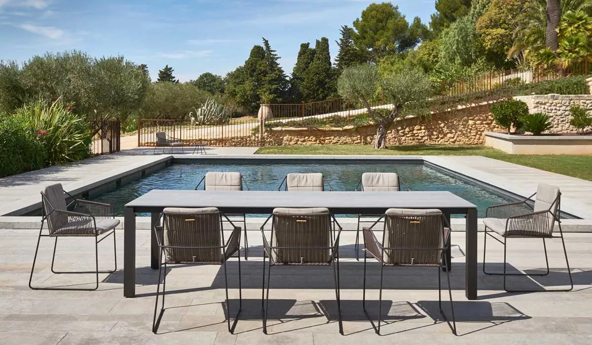 Oasiq Machar Essgruppe in Grau vor einem Pool auf einer Terrasse mit Olivenbäumen im Hintergrund, bestehend aus Tisch und sechs Stühlen.