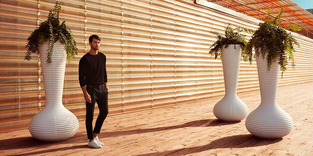 Mann in schwarzem Pullover neben drei weißen Vondom Bloom Pflanzkübeln auf Holzterrasse vor moderner Holzfassade.