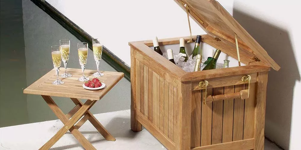 Barlow Tyrie Reims Kühlbox aus Teakholz mit geöffnetem Deckel, gefüllt mit Flaschen und Eis, daneben ein kleiner Tisch mit Gläsern.