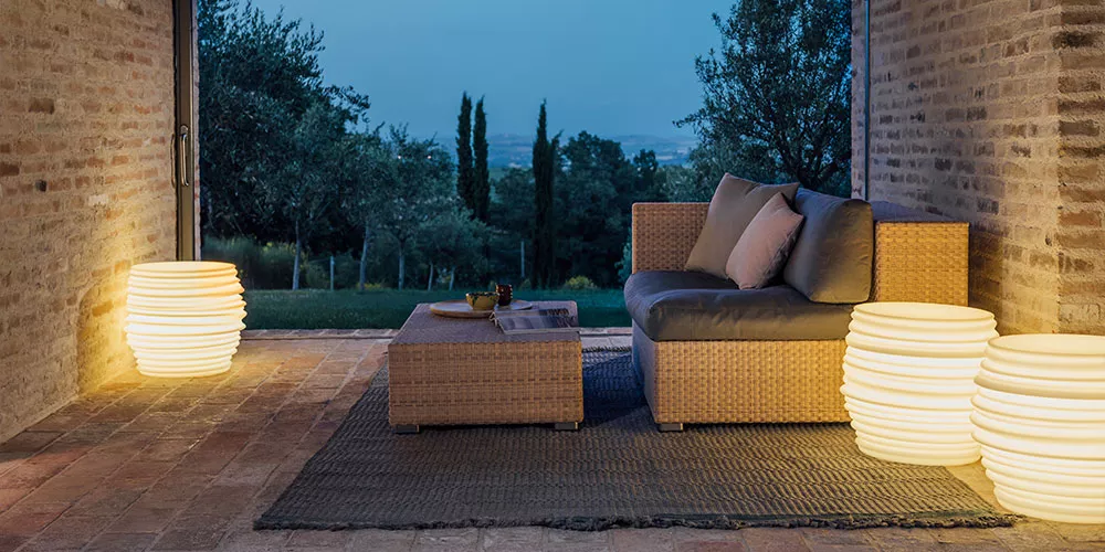 Dedon Babylon Leuchten beleuchten eine gemütliche Sitzecke im Freien bei Dämmerung mit einem Sofa und einem Couchtisch auf einem Teppich.