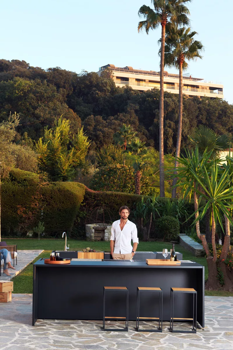 Röshults Kitchen Island Outdoor in Schwarz auf einer Terrasse mit Palmen im Hintergrund, stimmungsvolles Bild mit einem Mann im weißen Hemd.