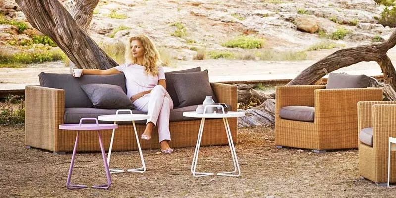 Cane-line Chester Loungegruppe in Beige und Grau auf sandigem Boden mit Bäumen im Hintergrund, Frau sitzt entspannt auf dem Sofa.