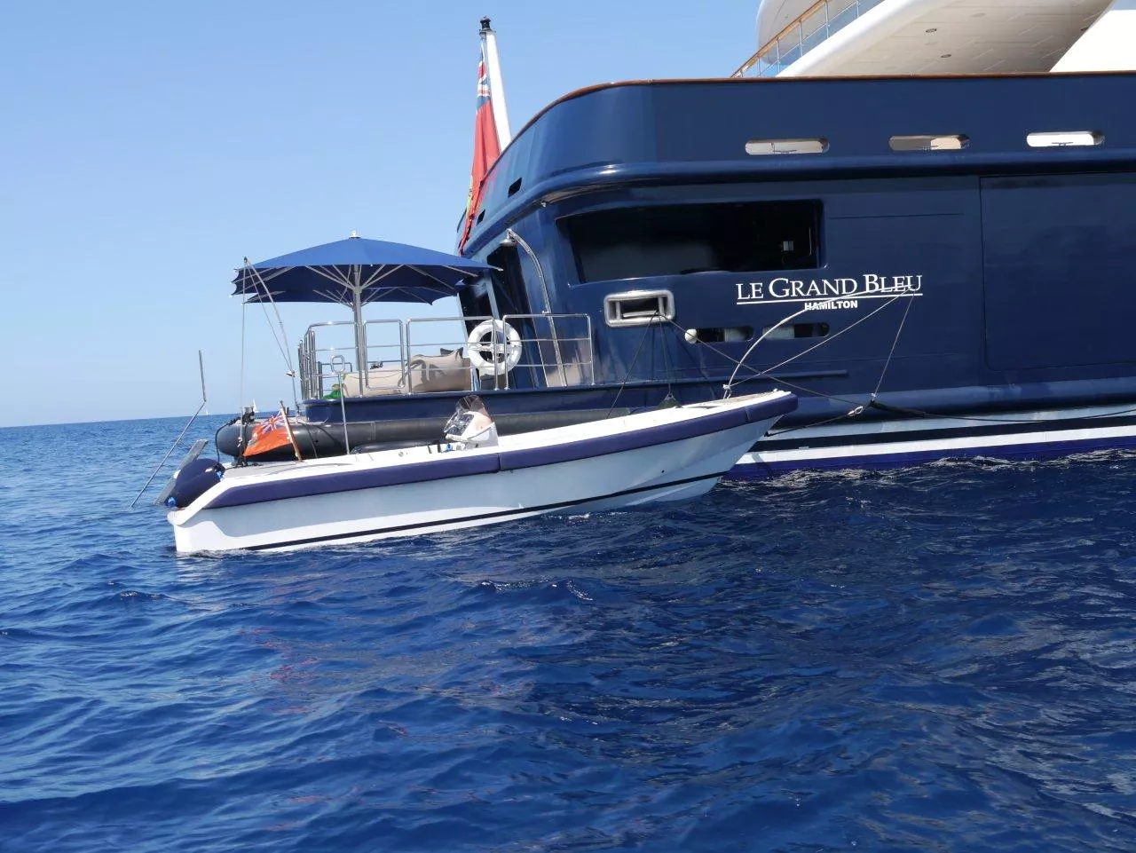 Ein kleines Motorboot mit Sonnenschirm ist neben einer großen Yacht namens Le Grand Bleu im Meer angedockt.