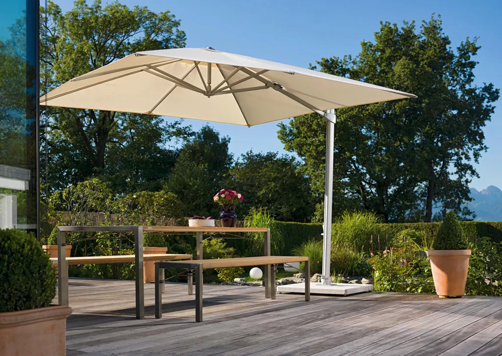 Freiarm-Sonnenschirm von Weishäupl in Beige auf einer Holzterrasse mit Gartenmöbeln und grüner Landschaft im Hintergrund.