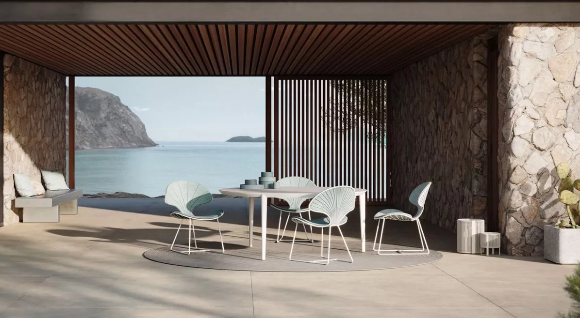 Outdoor-Moodbild mit Kettal Cala Stühlen in Hellblau und einem runden Tisch auf einer Terrasse mit Meerblick und Steinwänden.