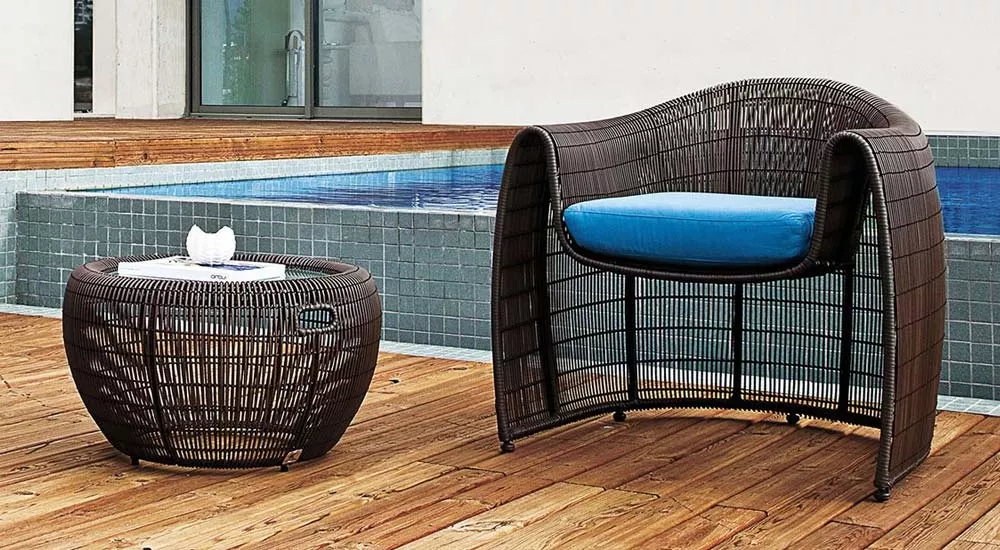 Brauner Kenneth Cobonpue Lulu Loungesessel mit blauem Kissen und passendem Beistelltisch auf Holzterrasse vor Pool.