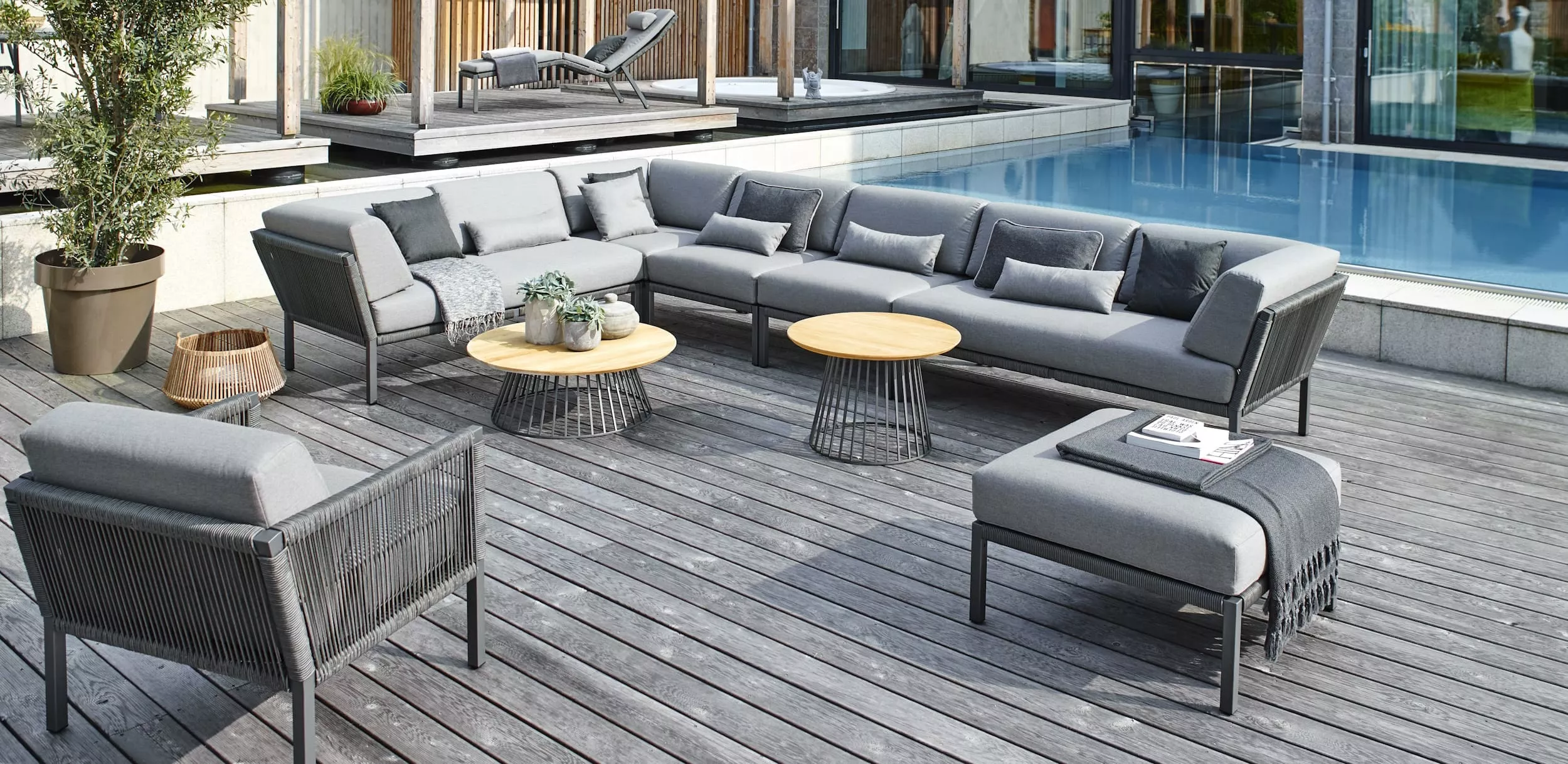 Solpuri Club Lounge Set in einem modernen Garten mit Pool und Holzterrasse im Hintergrund Mood Bild mit grauen Polstern und runden Beistelltischen