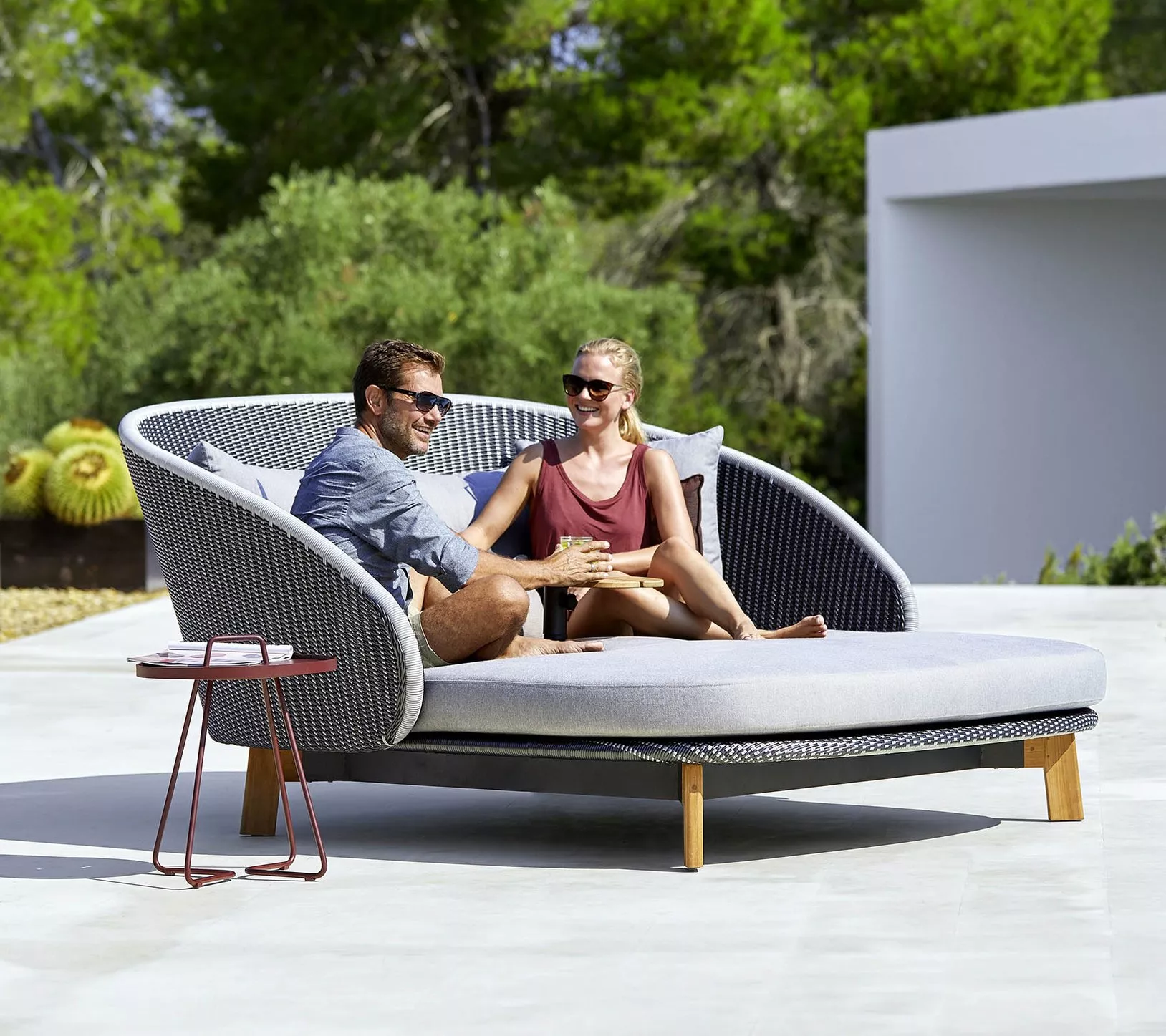 Cane-line Peacock Weave Daybed in Grau mit Teak-Beinen, 219 cm, auf einer Terrasse mit zwei Personen, umgeben von grüner Natur.