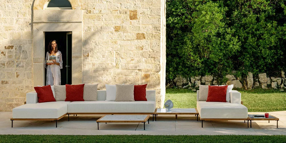 Talenti Kollektion Cleo Teak, weißes Outdoor-Sofa mit roten Kissen, vor einer Steinmauer im Garten.
