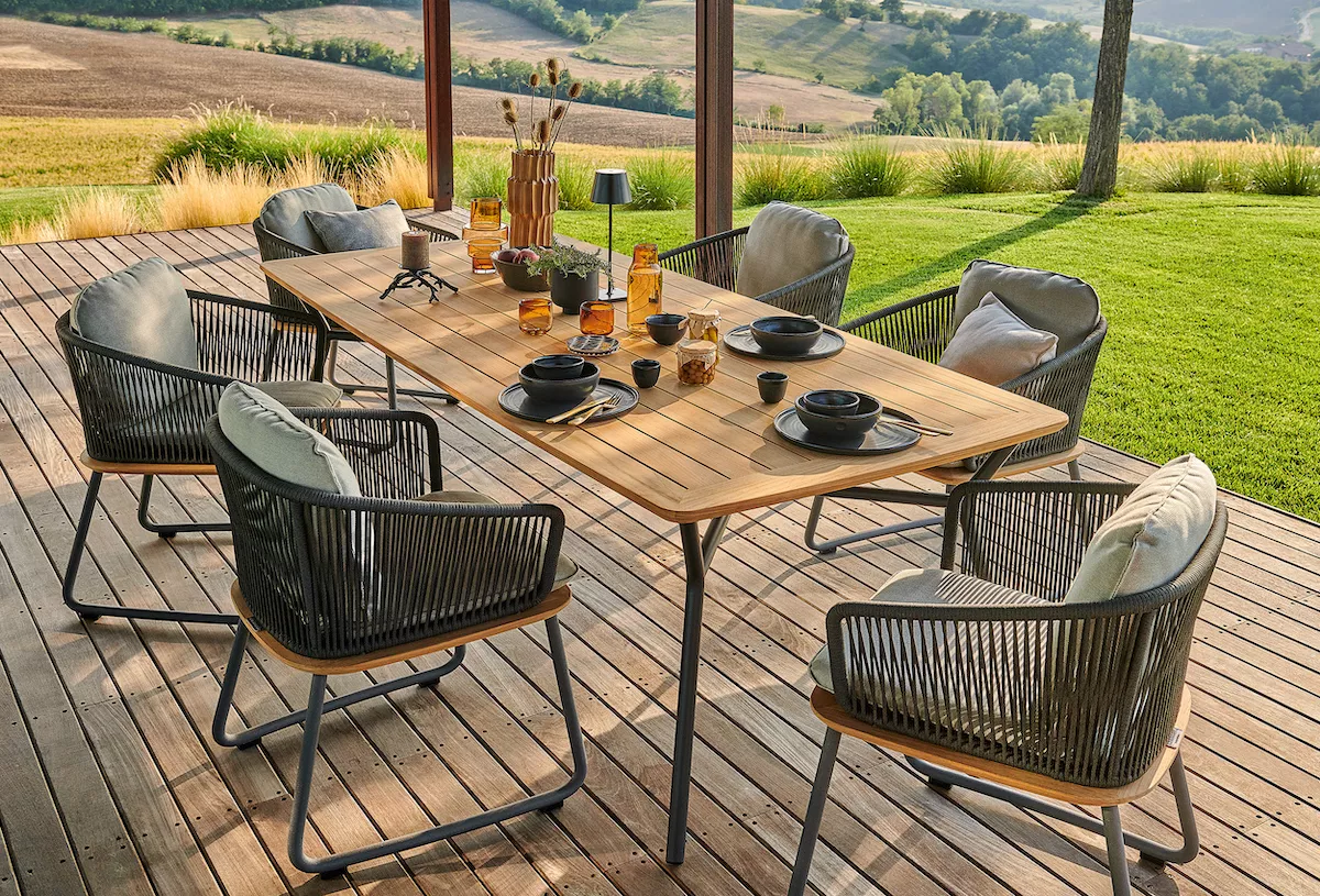 Weishäupl Denia Armlehnstuhl in Schwarz auf einer Terrasse mit Holztisch und Geschirr, umgeben von grüner Landschaft im Hintergrund.