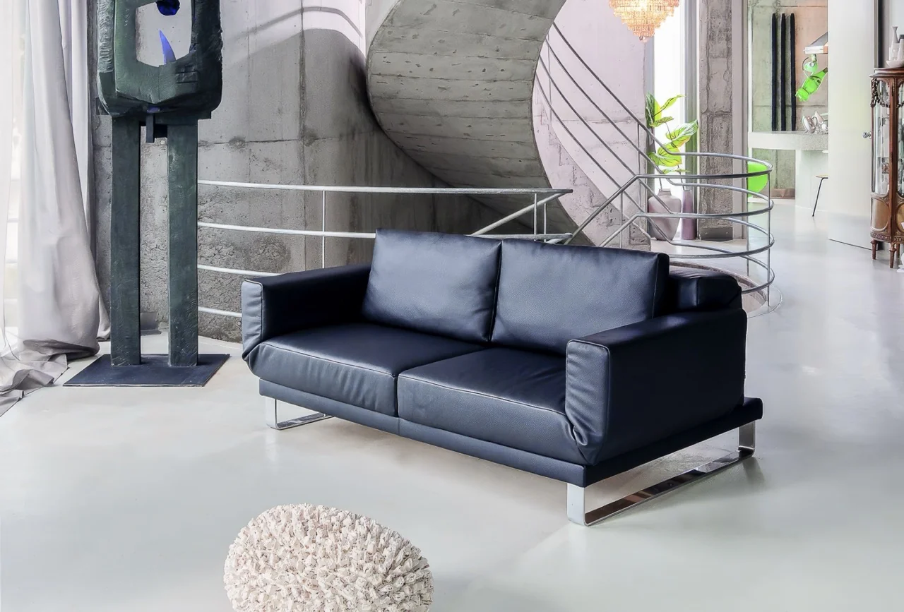 Schwarzes Franz Fertig RIGA-XL Sofa in modernem Loft mit Betonwänden und geschwungener Treppe im Hintergrund.