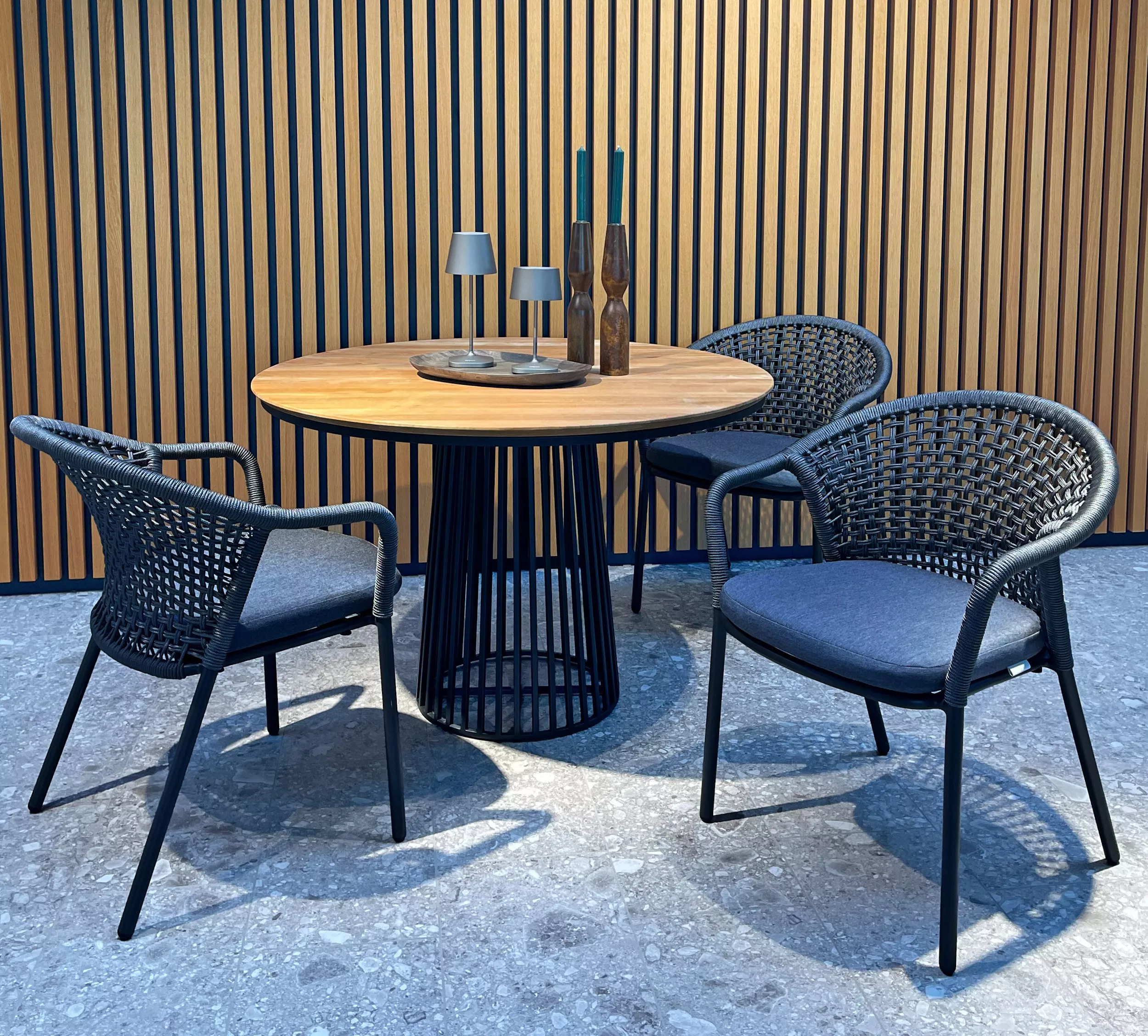 Solpuri Jooy Essgruppe mit rundem Holztisch und drei geflochtenen Stühlen in einem modernen Raum mit Holzlamellenwand und Dekoration.