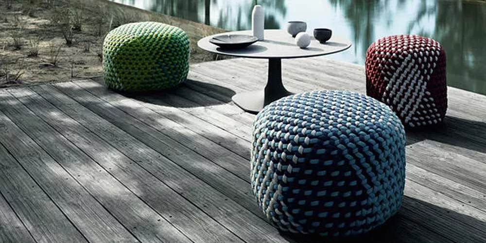 Drei BeB Italia Tramae Hocker in verschiedenen Farben stehen auf einer Holzterrasse neben einem runden Tisch mit Geschirr am Wasser