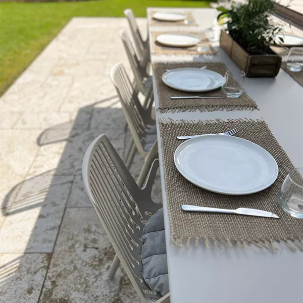 Gedeckter Gartentisch mit weißen Tellern und Gläsern auf beigem Tischläufer, umgeben von grauen Stühlen auf einer Steinplatte im Freien.