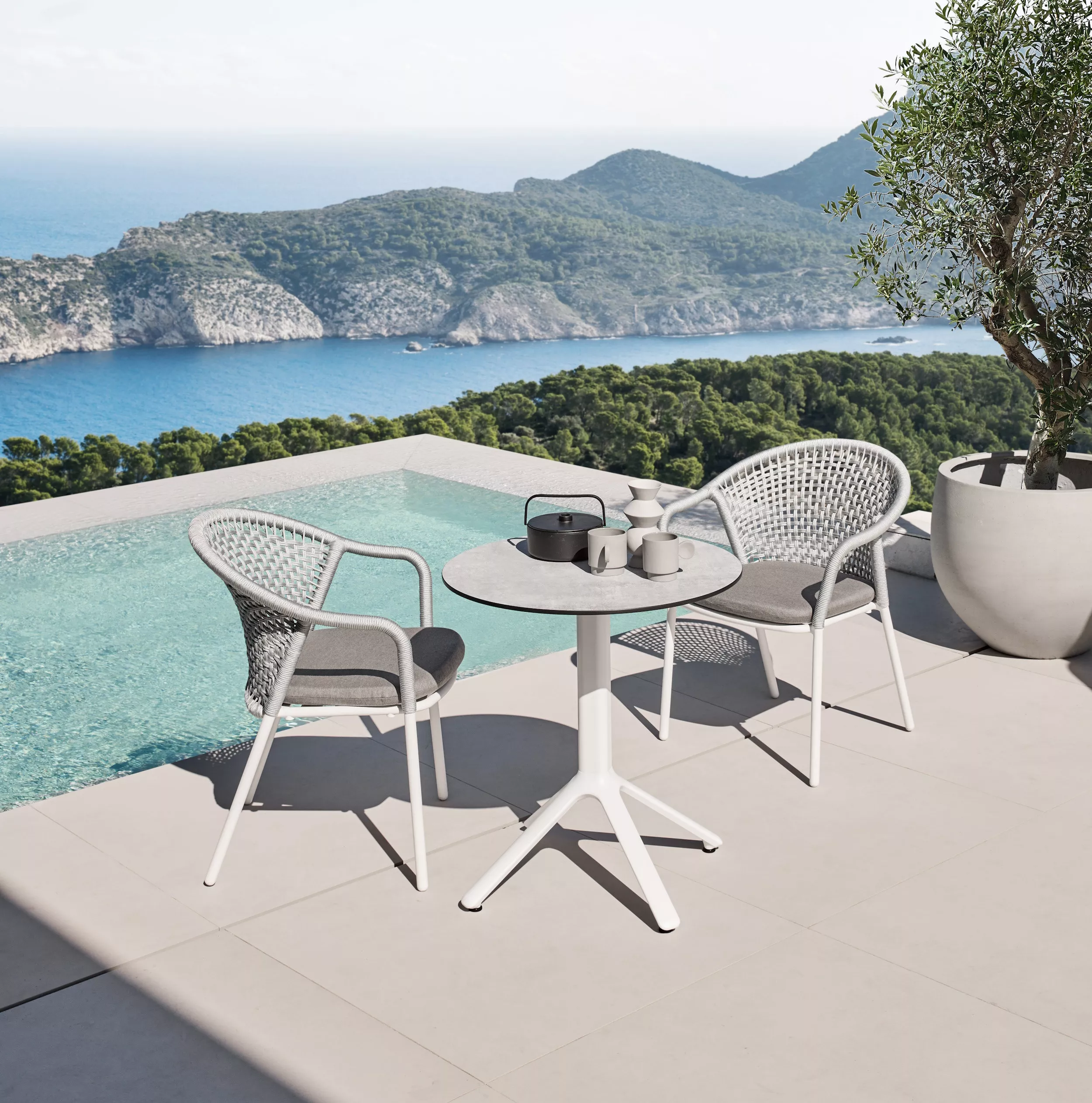 Zwei Solpuri Jooy Stühle und ein Tisch stehen auf einer Terrasse mit Blick auf das Meer und die Berge im Hintergrund Mood Bild