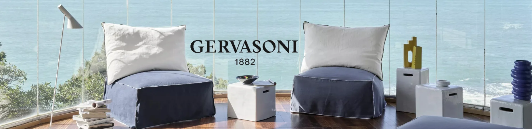 Zwei Gervasoni-Sessel in Blau und Weiß vor einer Glaswand mit Meerblick, umgeben von weißen Beistelltischen und Dekorationen.