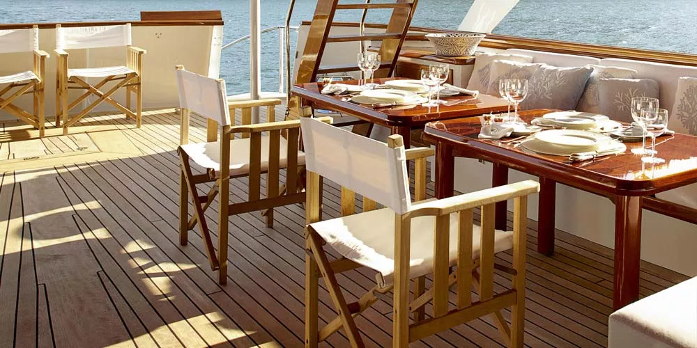 Barlow Tyrie Safari Essgruppe in Holzoptik auf einer Yacht, bestehend aus Tisch und Stühlen mit weißen Stoffbezügen, auf einem Deck.