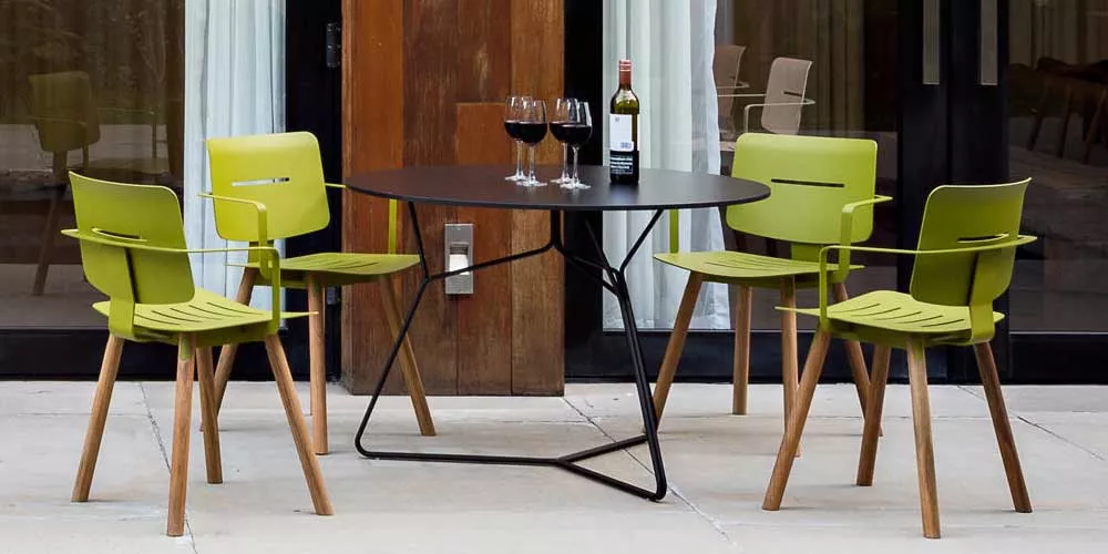 Oasiq Coco Essgruppe in Grün mit rundem Tisch, Gartenmöbel-Set auf Terrasse vor Glasfassade, zwei Weingläser und Flasche auf Tisch.