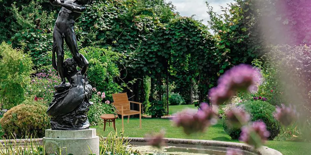 Gartenstimmung mit Bronzestatue im Vordergrund, umgeben von blühenden Pflanzen und einem Holzstuhl im Hintergrund.