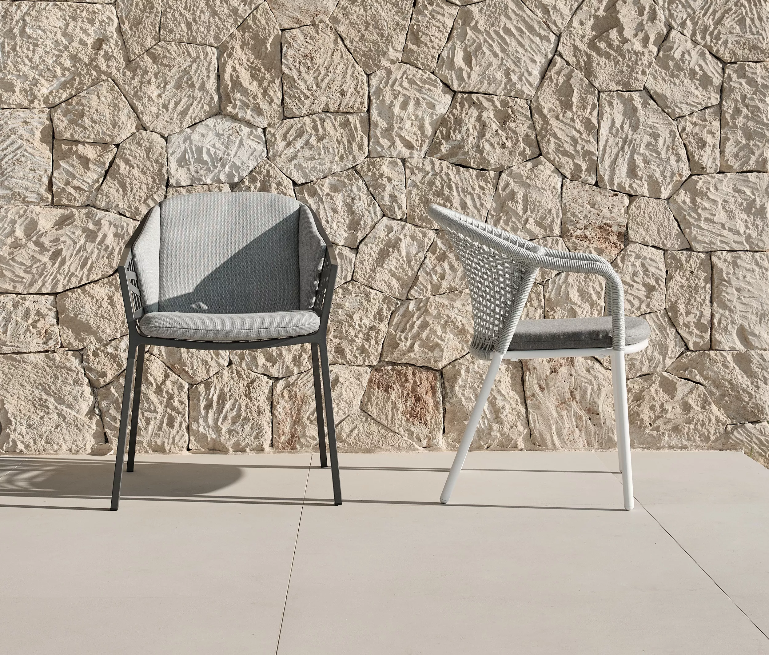 Zwei Solpuri Jooy Stühle in Grau und Weiß stehen vor einer Steinmauer auf einer Terrasse in der Sonne.