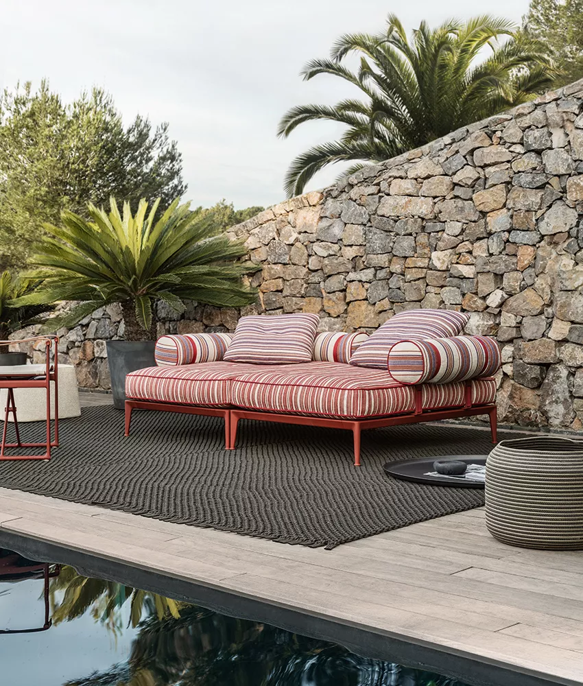B&B Italia Ribes Outdoor Lounge in einem Garten neben einem Pool mit Palmen und Steinmauer im Hintergrund.