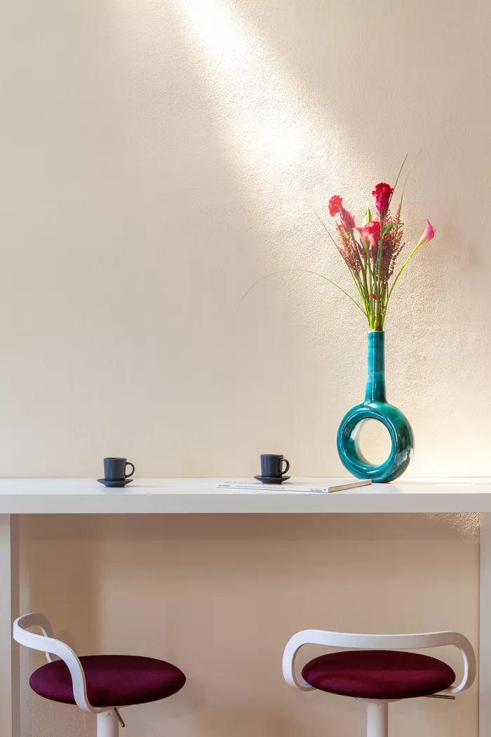 Zwei weiße Barhocker mit bordeauxroten Sitzflächen vor einem schlichten weißen Tisch, darauf eine türkisfarbene Vase mit Blumen, in einem hellen Raum.