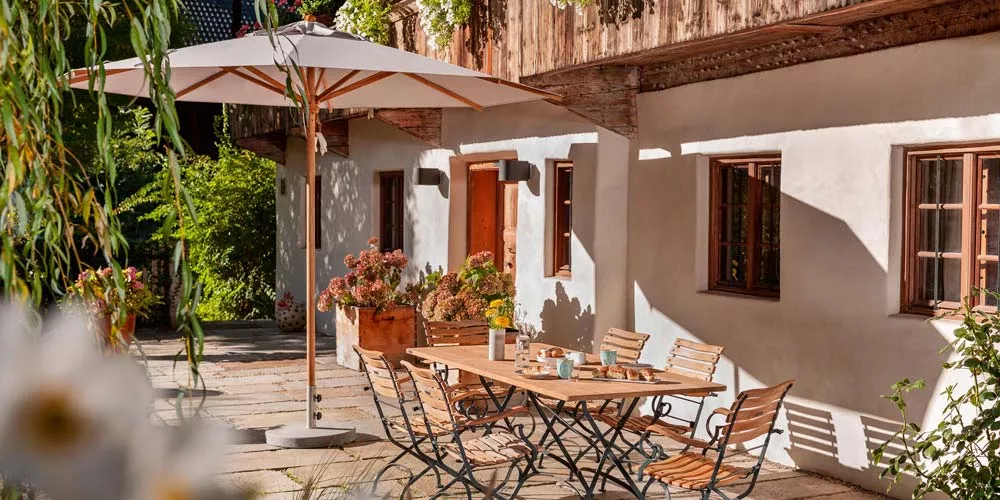 Weishäupl Teakschirm in Beige auf einer sonnigen Terrasse mit Holzmöbeln und blühenden Pflanzen im Hintergrund.