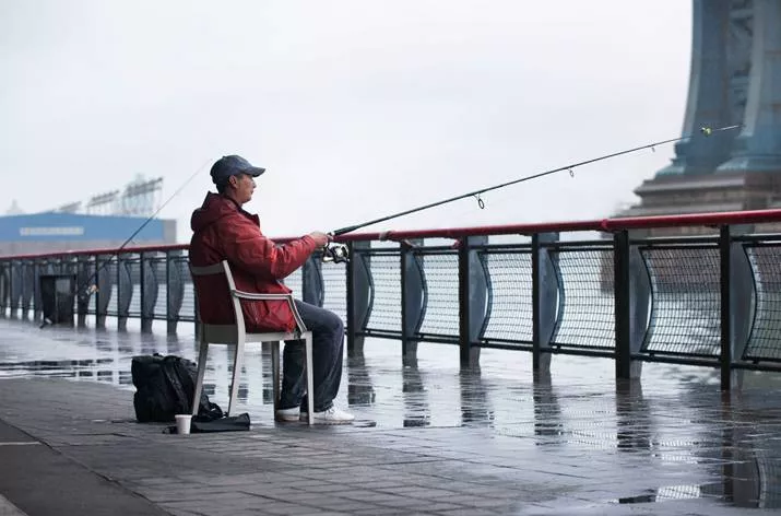 Ein Mann sitzt auf einem Dedon Play Essstuhl und angelt an einem regnerischen Tag an einem Geländer mit Blick auf eine Brücke.