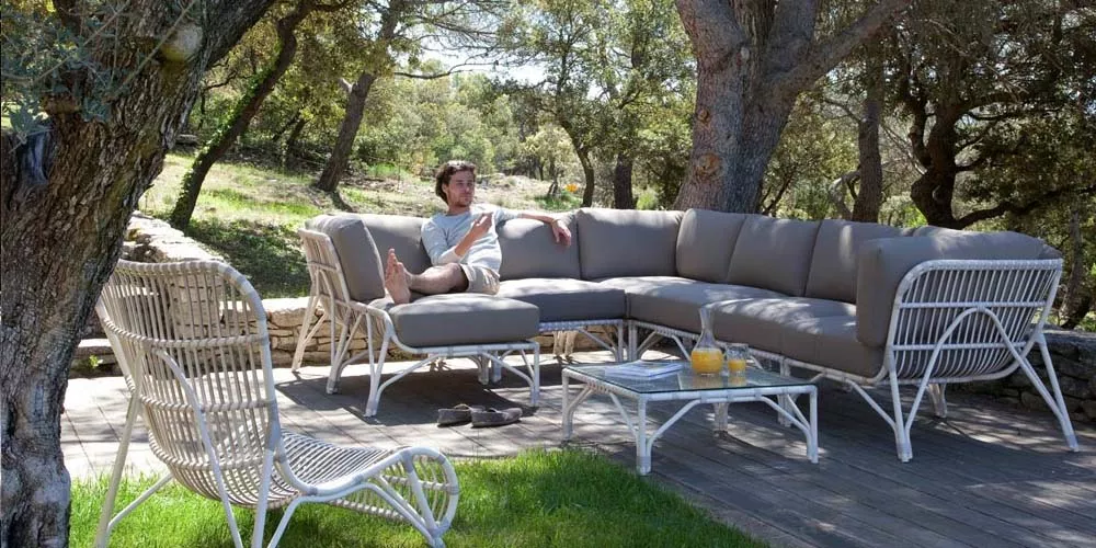 Vincent Sheppard Lucy Lounge-Set in Beige auf Holzterrasse im Garten, bestehend aus Sofa, Tisch und Stuhl, umgeben von Bäumen.