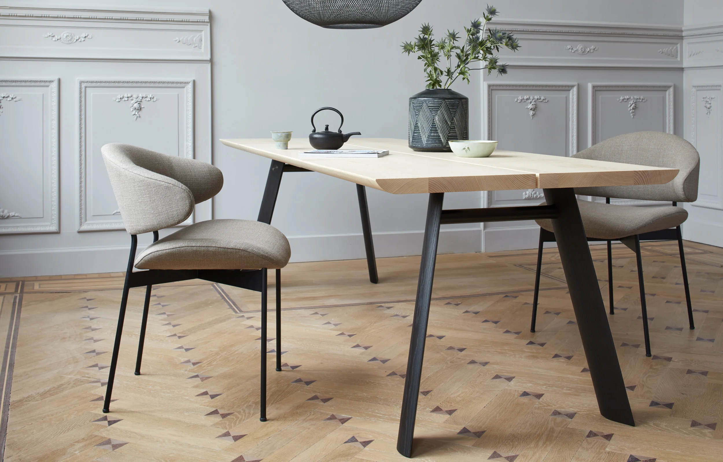 LUZ Stuhl von more in Beige, vor einem Holztisch in einem eleganten Raum mit Parkettboden und dekorativen Wandpaneelen.