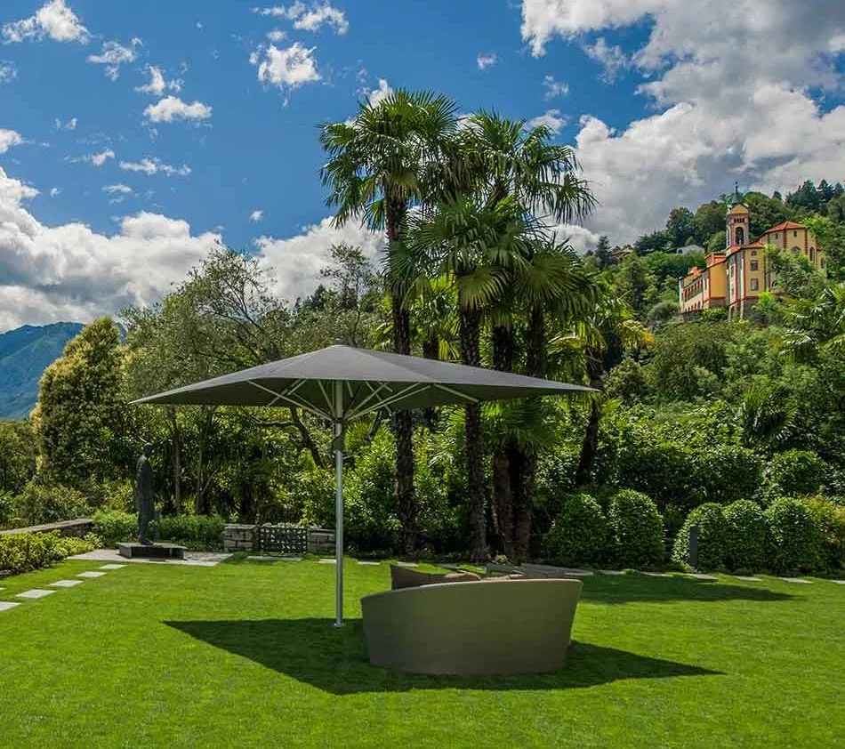 Sonnenschirm Fortello von ABB in Grau auf einer grünen Wiese vor einer Villa, umgeben von Palmen und Bergen, bei sonnigem Wetter.