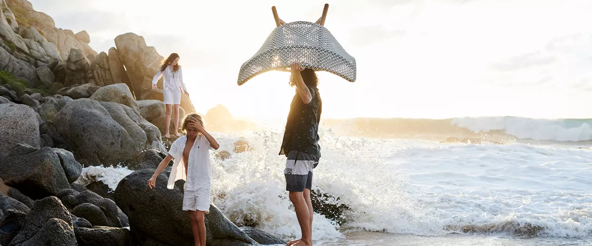 Personen am Strand mit einem Rattansessel von Villa Schmidt, im Hintergrund Felsen und Meer, stimmungsvolle Abendsonne.