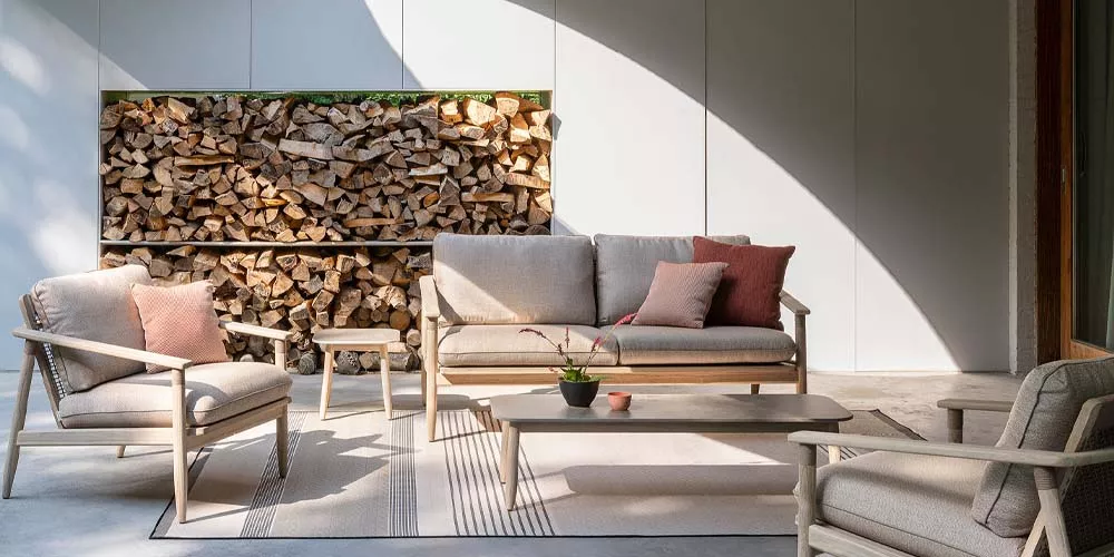 Vincent Sheppard David Lounge-Set in Beige auf einer Terrasse mit Holzstapel im Hintergrund, bestehend aus Sofa, Sesseln und Tisch.