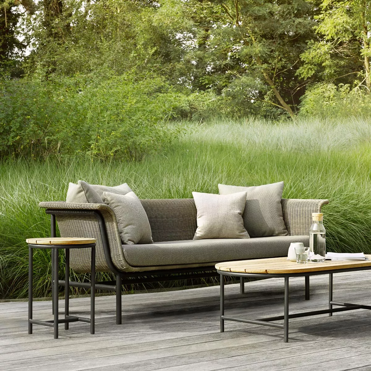 Vincent Sheppard Wicked Loungegruppe in Grau auf Holzterrasse vor grüner Gartenkulisse, bestehend aus Sofa und Beistelltischen.