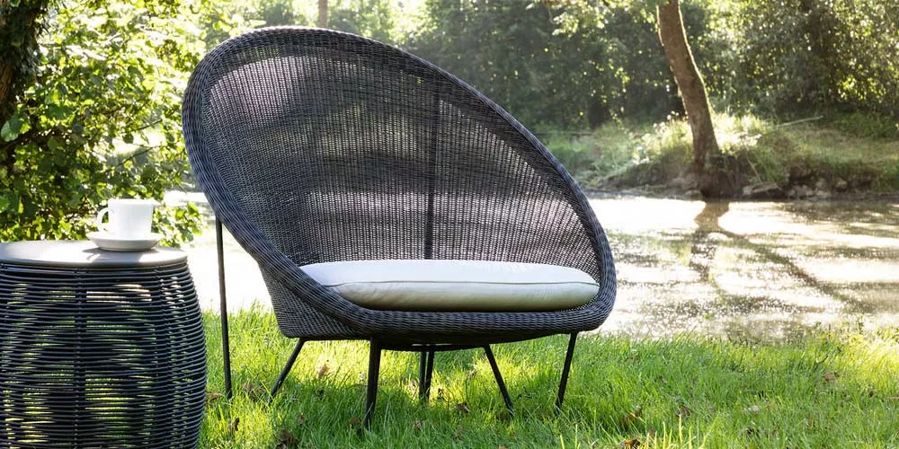 Vincent Sheppard Gipsy Sessel in Schwarz auf grünem Rasen vor einem Teich, mit weißem Kissen und Beistelltisch mit Kaffeetasse.